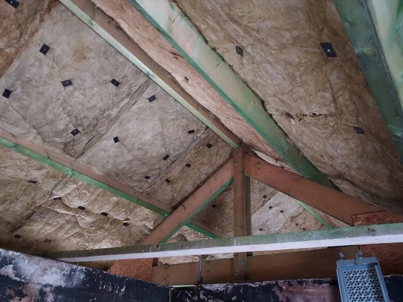 `Isolation de toiture par l'intérieur avec une double couche de laine minérale. Vue sur la charpente en bois, avec des panneaux d'isolation fixés à l'aide de dispositifs de maintien. Travaux réalisés pour améliorer l'efficacité thermique du bâtiment. Structure solide avec poutres en bois apparentes, dans un contexte de rénovation énergétique.`