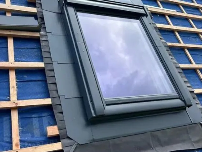`Pose d'une fenêtre de toit de type Velux avec un raccord d'étanchéité sur une toiture en rénovation. La sous-toiture bleue et le lattage en bois sont visibles, indiquant des travaux en cours pour l'installation de la couverture finale. Fenêtre parfaitement intégrée pour garantir l'isolation thermique et l'étanchéité à l'eau.`